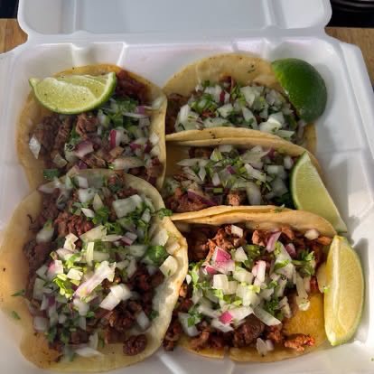 Four street tacos topped with chopped onion and cilantro, with lime wedges
